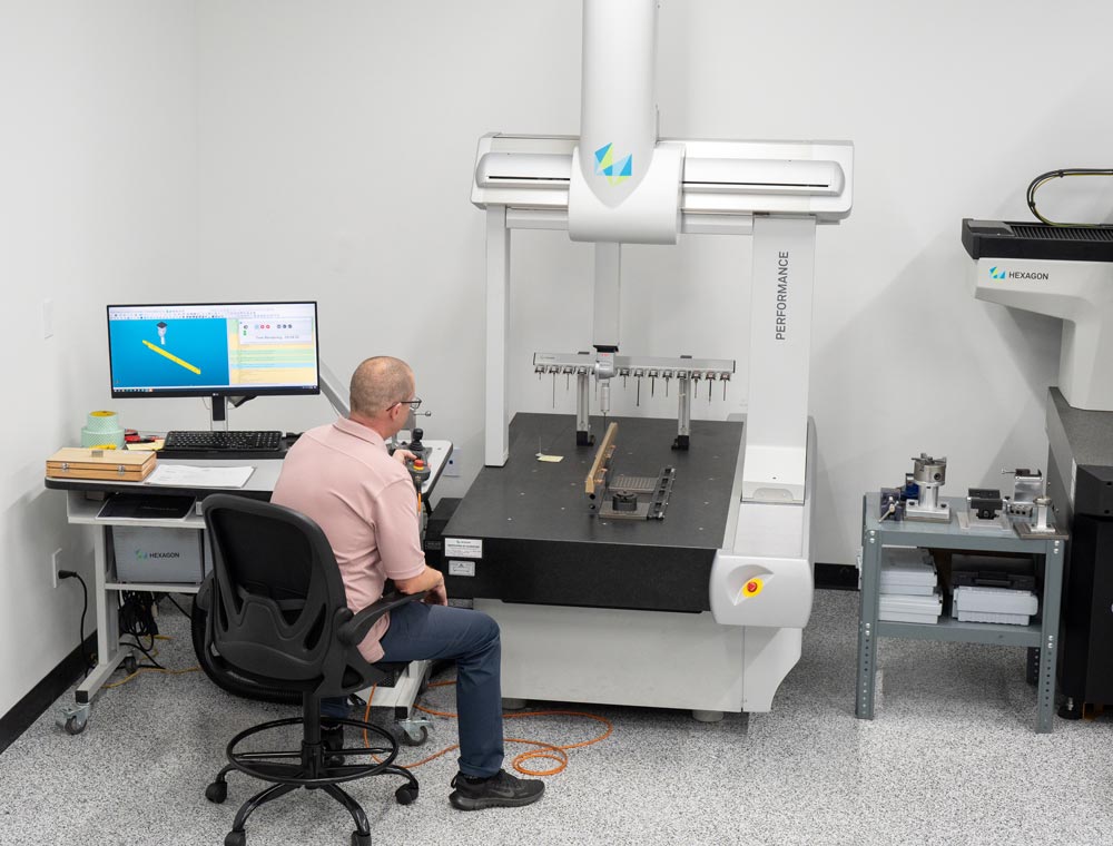 A man sits at a workstation operating a coordinate measuring machine (CMM) in a lab setting.