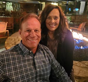 A smiling man and woman in casual attire sit by an outdoor fire pit at a cozy evening gathering.