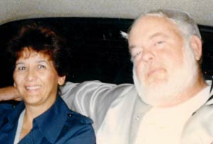 A smiling woman with short dark hair and a man with a white beard sit closely together, the man's arm around the woman.