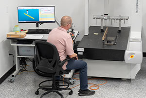 A man operates a coordinate measuring machine at a workstation with a computer monitor displaying measurement data.