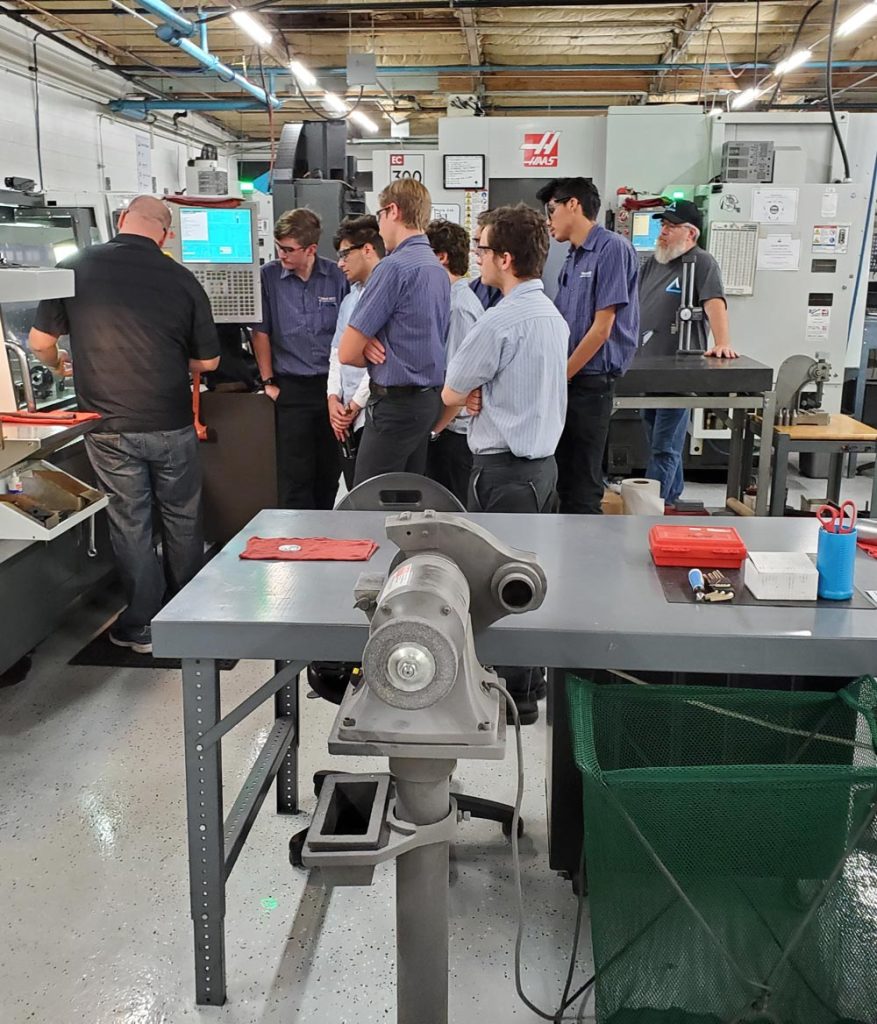 A group of people watches a man operate a machine in an industrial workshop setting with equipment in the foreground.