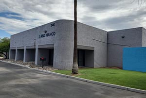 A modern gray building with "The Land Group" sign, fronted by a palm tree, lawn, and parking lot under a cloudy sky.