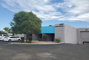 A grey building with a blue awning, labeled "5127," with a tree, bushes, and parked cars in front.