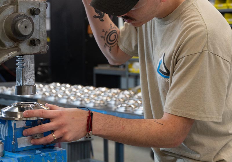 Person operating a milling machine, focusing on a metal part, with a table of round metal objects in the background.