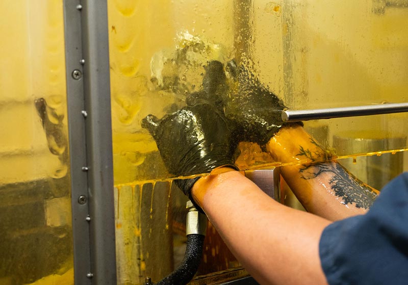 A person wearing black gloves works on machinery covered with a yellow liquid, with a tattoo visible on their arm.