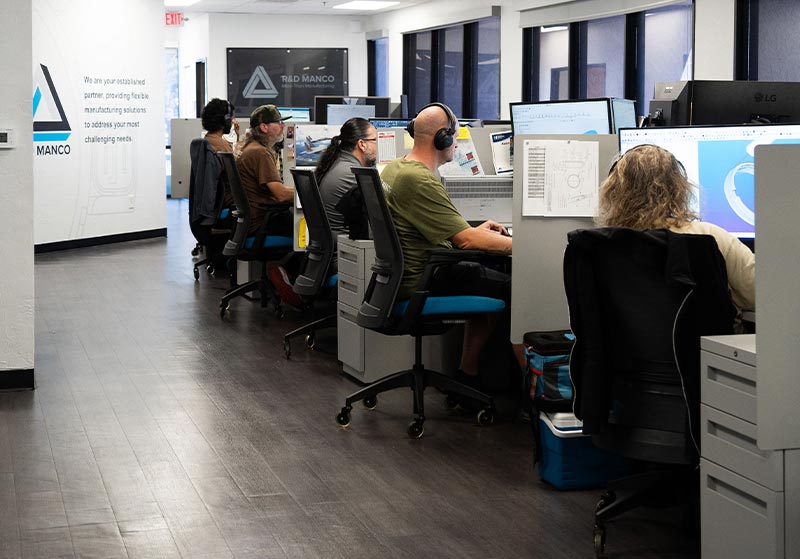 People working at computer desks in an office, each with a headset, large screens, and various technical drawings around.